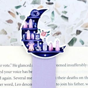 May include: A magnetic bookmark featuring a crescent moon shape with shelves holding crystals, bottles, and flowers, attached to a solid light purple rectangular base. The text JustAddJewelry is printed in a white bubble shape at the bottom.