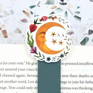 May include: A magnetic bookmark featuring a circular top with a yellow crescent moon illustration surrounded by colorful flowers and leaves, attached to a dark teal rectangular base with the white text JustAddJewelry printed on the bottom.