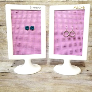 May include: Two white earring display stands with pink mesh grids. The stand on the left has the name "Emma" written on it and has two teal blue stud earrings. The stand on the right has the name "Abbie" written on it and has two silver hoop earrings.