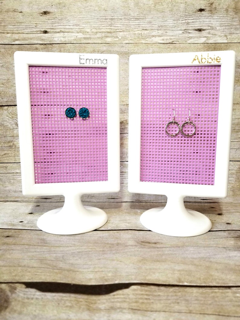 May include: Two white earring display stands with pink mesh grids. The stand on the left has the name "Emma" written on it and has two teal blue stud earrings. The stand on the right has the name "Abbie" written on it and has two silver hoop earrings.