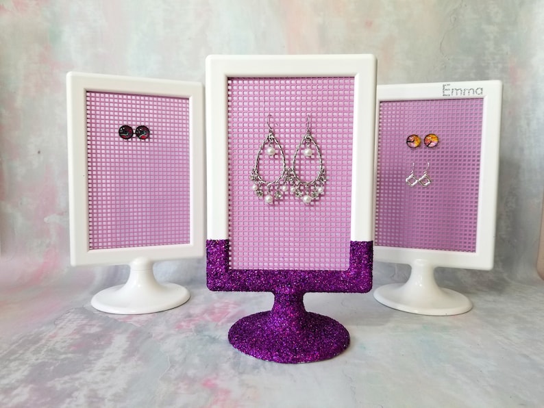 May include: Three white framed earring display stands with pink mesh backing. The center stand has a purple glitter base and displays a pair of silver dangle earrings with white pearls. The stands on either side have white bases and display pairs of stud earrings.