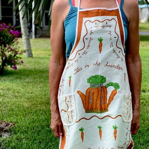 Könnte beinhalten: Eine weiße Schürze mit einem orange-grünen Design, das eine Teekanne und Karotten zeigt. Die Schürze trägt den Text "Tea in the Garden".