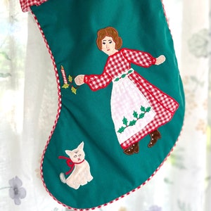 May include: A festive Christmas stocking with a red top and a green body. The top is trimmed with red and white gingham. The stocking is decorated with embroidered images of a woman, a cat, and a candle.