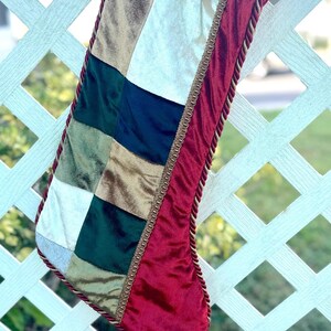 May include: A red velvet Christmas stocking with a patchwork design of green, gold, and white velvet. The stocking has a gold cord trim and a bell at the end.
