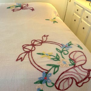 May include: A pale pink bedspread with a ribbed texture, featuring embroidered floral and ribbon designs in red, green, blue, and yellow. The bedspread is covering a bed.