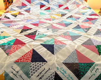 May include: A white quilt with a patchwork design of colorful triangles. The quilt is signed with names in black ink, including "James A. Hanno", "Stilla Pachend", and "Grampa Miller".
