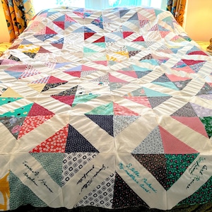 Puede incluir: Una colcha blanca con un diseño de patchwork de triángulos de colores. La colcha está firmada con nombres en tinta negra, incluyendo "James A. Hanno", "Stilla Pachend" y "Grampa Miller".
