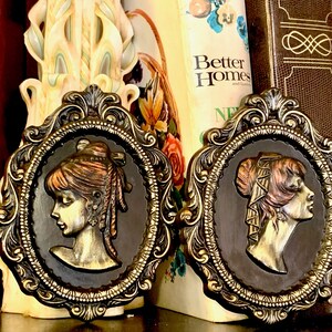 Könnte beinhalten: Zwei verzierte goldfarbene Rahmen mit dunkelbraunen Hintergründen, die jeweils ein erhabenes Kamee eines Frauenprofils im Vintage-Stil zeigen. Die Frauen haben braunes Haar und tragen aufwendige Frisuren.