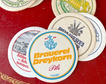 May include: A collection of round, white paper coasters with various designs and text. The coasters feature logos and text in German, including "Brauerei Dreykorn Pils" and "Lowenbrau".