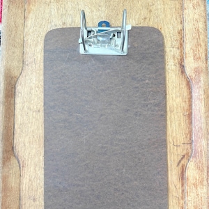 Può includere: Un portablocco marrone con clip in metallo, su sfondo in legno. Il portablocco ha una forma rettangolare con angoli arrotondati. La clip in metallo è argentata e tiene fermo il foglio.