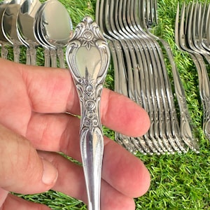 May include: Close-up of a silver fork with ornate floral detailing on the handle. The fork is part of a set of silverware, with other forks and spoons visible in the background. The silverware is set against a green background.
