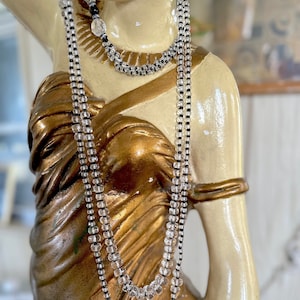 May include: A long, layered necklace with clear glass beads and black beads. The necklace is worn by a mannequin with a gold-painted torso.