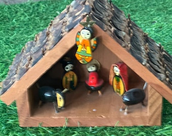 May include: A wooden nativity scene with a brown roof and a collection of painted figurines. The figurines include an angel, a woman in a red head covering, and other figures. The scene is set on a green surface.