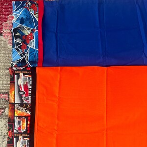 May include: A collection of colorful pillowcases. One pillowcase features a Spider-Man design with a blue background. Another is solid blue, and a third is bright orange. The last pillowcase has a firefighter theme.