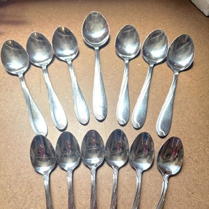 Könnte beinhalten: Ein Set mit 15 silbernen Teelöffeln mit einem dekorativen Wirbelmuster am Griff. Die Löffel sind in zwei Reihen auf einer Holzoberfläche angeordnet.