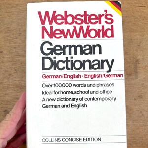 Op de afbeelding: Een wit boek met de titel "Webster's NewWorld German Dictionary" met rode en zwarte tekst. Het boek is een Collins Concise Edition en bevat Duitse/Engelse en Engelse/Duitse vertalingen. De omslag toont ook de kleuren van de Duitse vlag.