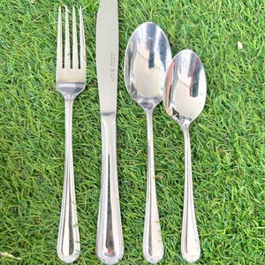 May include: A collection of four silver-coloured cutlery items: a fork, a knife, and two spoons. The items are displayed on green grass. The knife has a serrated edge and a decorative handle. The spoons are of different sizes.