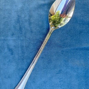 Könnte beinhalten: Ein silberner Löffel mit einem dekorativen Griff. Der Löffel liegt auf einer blauen Stoffoberfläche.