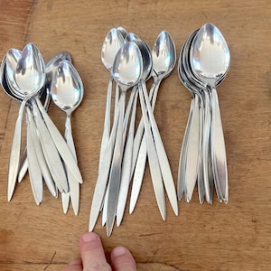 May include: A collection of shiny, silver spoons with elongated handles. The spoons are arranged in several groups on a wooden surface. The spoons have a classic, elegant design, reflecting light. The image showcases a set of silverware.