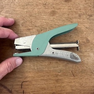 May include: A vintage stapler with a silver body and a mint green handle. The stapler is made of metal and has a lever-style design. The image shows the stapler on a wooden surface.