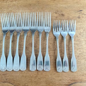 May include: A collection of silver forks arranged on a wooden surface. The forks have a classic design with tines and decorative handles. The arrangement includes both large and smaller forks, suitable for various courses.