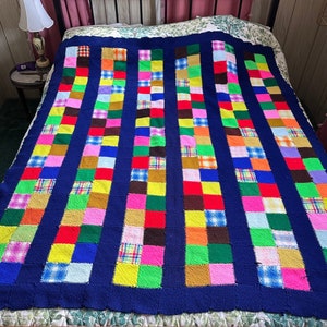 May include: A handmade patchwork quilt with a navy blue border and colorful squares. The squares are arranged in vertical rows and feature various patterns and colors, including red, yellow, green, and plaid. The quilt is displayed on a bed.