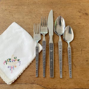 Könnte beinhalten: Ein Vintage-Besteckset mit einem dekorativen Blumendesign an den Griffen, einschließlich Gabeln, einem Messer und Löffeln. Ein weißes, besticktes Taschentuch mit einem Blumengestaltung ist ebenfalls vorhanden. Die Artikel sind auf einer Holzoberfläche angeordnet.