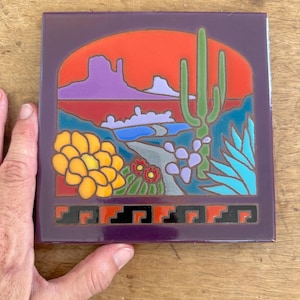 May include: Square ceramic tile with a desert landscape design. The tile features a purple border, an orange sunset, and a depiction of cacti, mountains, and desert flora in various colors. The bottom border has a geometric pattern.