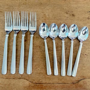 May include: A set of four silver-toned metal forks with four tines each and five silver-toned metal spoons with oval bowls. The flatware features smooth, plain handles and a polished finish, arranged in two parallel rows on a wooden surface.