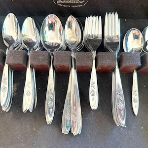 May include: A set of silver-colored flatware, including spoons, forks, and serving utensils, arranged in a black storage case. The handles feature a floral rose design. The utensils are held in place by brown and black foam inserts.