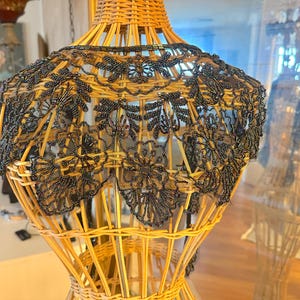 May include: A black beaded collar with floral designs is displayed on a wicker dress form. The collar has a scalloped edge and intricate beadwork. The dress form is made of woven wicker and has a natural, light brown color.