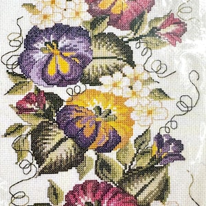 Op de afbeelding: Borduurwerk met kruissteek van een bloemstuk met paarse, gele en roze bloemen met witte bloesems en groene bladeren. De bloemen zijn verticaal gerangschikt met een rankmotief.