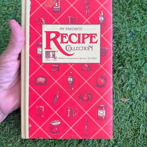 May include: A vintage cookbook titled "My Favorite Recipe Collection" with a red cover featuring food-related illustrations. The book's title is in a cream-colored banner. The book is held by a hand against a green grass background.