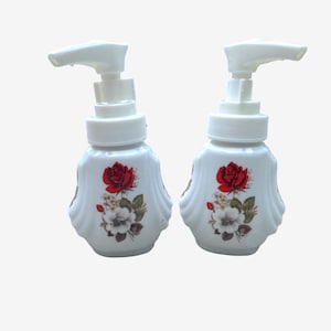 May include: Two white soap dispensers with pump mechanisms, featuring a decorative floral print on the front. Each bottle has a rounded, ribbed body design and displays a central graphic of a vibrant red rose above smaller white flowers.