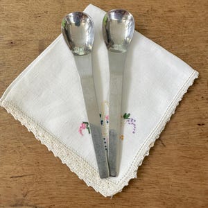 May include: Two silver serving spoons with oval bowls and long, flat handles rest on a white, embroidered napkin with a scalloped lace edge. The spoons are reflective, and the napkin has floral embroidery.