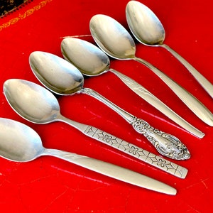 May include: Six silver spoons with different designs on a red surface. The spoons have a shiny finish and are arranged in a row.