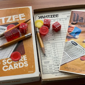 May include: A close-up of a Yahtzee game box with red dice, pencils, and score cards. The box is orange and brown with the words "Original Yahtzee" and "80 Score Cards".