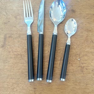 May include: A stainless steel flatware set including a fork, knife, and two spoons. The utensils have black handles with a silver band at the base. The set is arranged on a wooden surface.