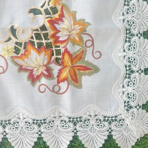 May include: White tablecloth with embroidered autumn leaves in shades of orange, yellow, and red. The tablecloth has a delicate, scalloped lace border. The design features a green trellis pattern.