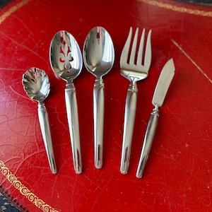 May include: Five pieces of cutlery on a red surface. The cutlery includes a fork, a butter knife, a spoon, a slotted spoon, and a small spoon with a scalloped edge.