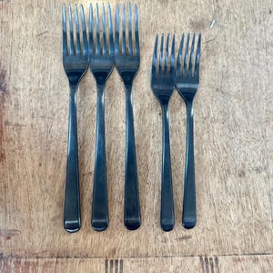 May include: Six stainless steel forks with a dark finish are arranged on a wooden surface. The forks are of two different sizes, with the larger forks having four tines and the smaller forks having three tines. The forks are evenly spaced.