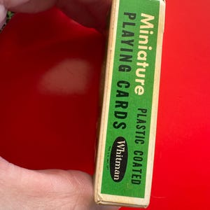 May include: A miniature deck of Whitman playing cards in a green and white box. The box is plastic coated and the text on the box reads "Miniature Playing Cards". The box is held by a person's hand.