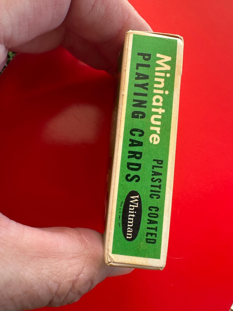May include: A miniature deck of Whitman playing cards in a green and white box. The box is plastic coated and the text on the box reads "Miniature Playing Cards". The box is held by a person's hand.