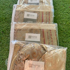Op de afbeelding: Verschillende Home Collection valances in doorzichtige plastic verpakking. De valances hebben een goud- en beige kleurenschema met een patroon. Elk pakket heeft een label met de tekst "home collection" en "valance".