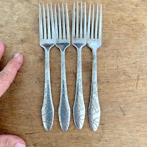 Puede incluir: Un juego de cuatro tenedores de metal plateado con un patrón decorativo en relieve en los mangos. El diseño incluye racimos de uvas grabados, zarcillos de vid y líneas curvas que se extienden a lo largo de cada mango hasta la base.