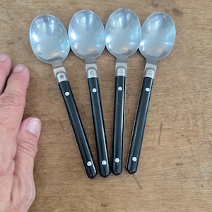 May include: A set of four spoons with polished silver-tone metal bowls and black handles. Each handle is secured with two small silver-tone rivets. The spoons are arranged side-by-side, featuring a classic design with a rounded bowl and a straight, slender handle that tapers slightly toward the end.