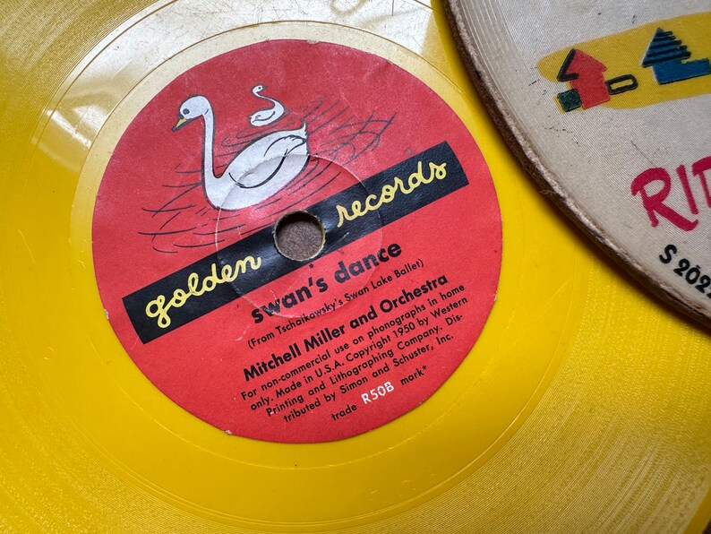 May include: A yellow vinyl record with a red label featuring a white swan and the text "golden records swan's dance (From Tschaikowsky's Swan Lake Ballet) Mitchell Miller and Orchestra For non-commercial use on phonographs in home only. Made in U.S.A. Copyright 1950 by Western Printing and Lithographing Company. Distributed by Simon and Schuster, Inc. trade R50B mark"