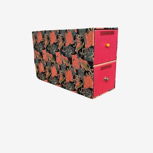 May include: A rectangular storage unit with two stacked drawers featuring bright pink fronts and a black patterned exterior. The top drawer has a small yellow knob and a label reading PAINT BRUSHES, while the bottom drawer has a silver knob and a label reading SCRAP BOOK.