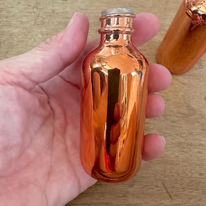 May include: A small, copper-colored glass bottle with a silver-colored cap. The bottle has a reflective, metallic finish. The bottle is held in a hand. Another copper-colored object is visible in the background.