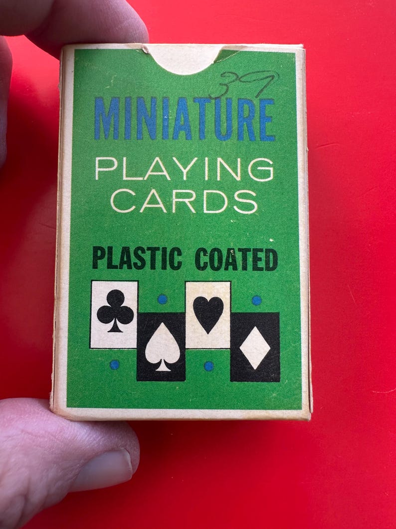 May include: A miniature deck of playing cards in a green box with blue and white text. The box features the words "MINIATURE PLAYING CARDS PLASTIC COATED" and card suit symbols. The number "39" is written in the top right corner.